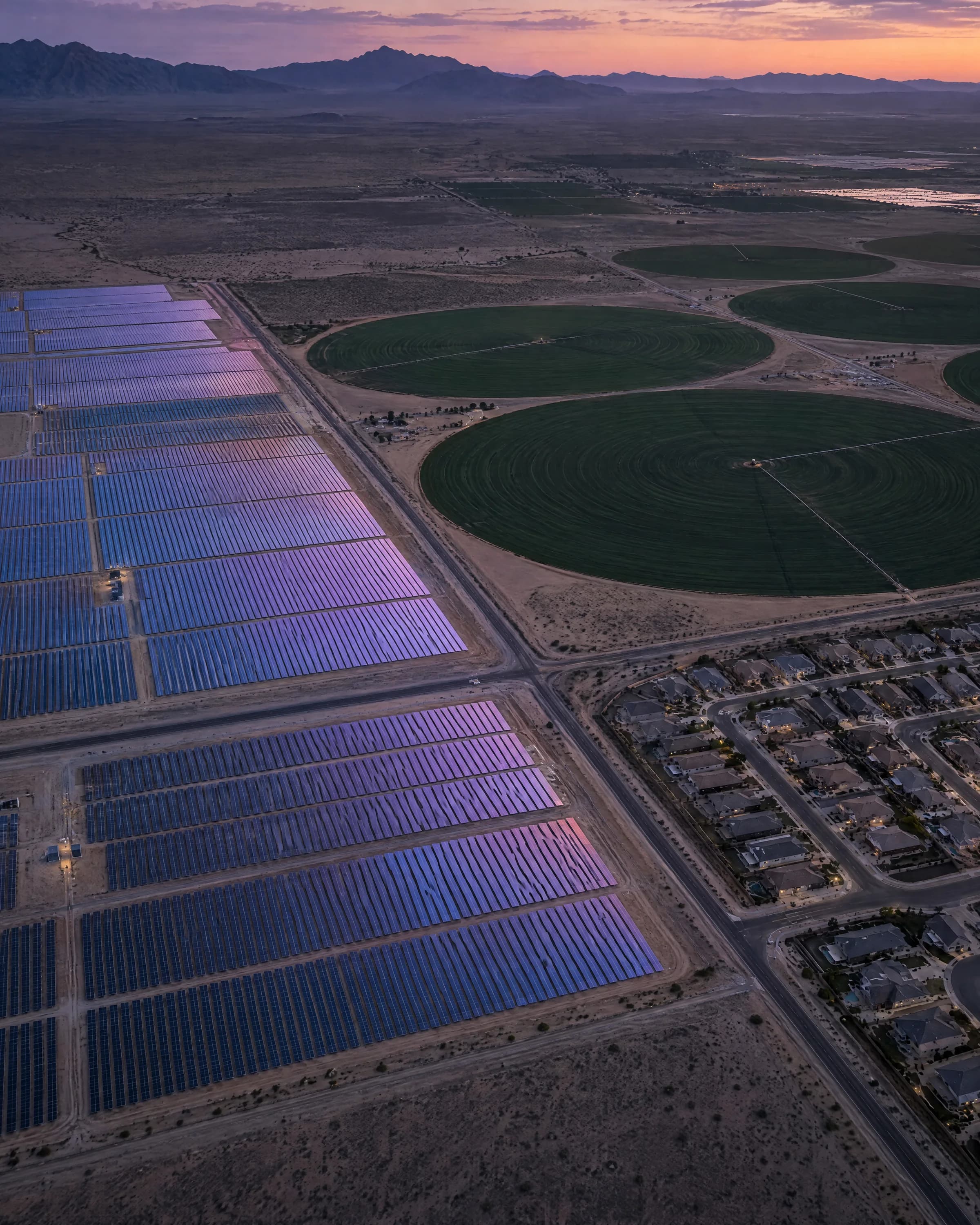 Aerial view of a desert solar farm, circular irrigated fields and a housing grid at dusk.